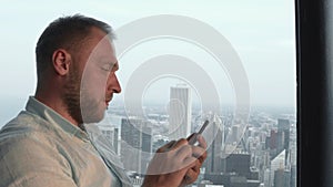 Young handsome man standing near a window in skyscraper and using smartphone. Panorama of Chicago, America.