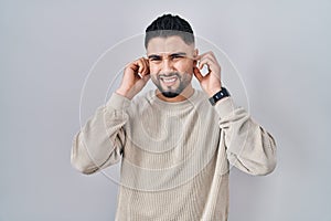 Young handsome man standing over isolated background covering ears with fingers with annoyed expression for the noise of loud