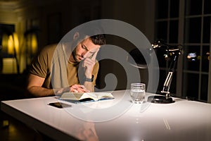 Young handsome man studying at home, reading a book at night