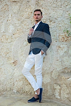 Young handsome man in a suit and white trousers at standing near a stone wall