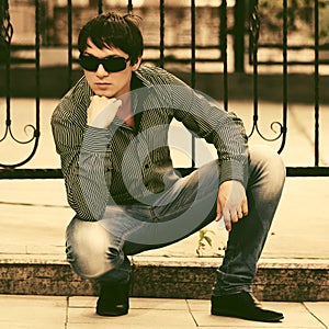 Young handsome man in sunglasses sitting on sidewalk
