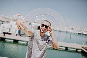 Young handsome man in sunglasses take selfie mobile phone on summer street with yacht harbor on background. Travel concept