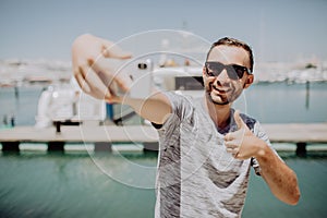 Young handsome man in sunglasses take selfie phone with thumbs up on yacht harbor on background. Travel concept