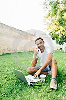 Young handsome man talking on the phone while working on laptop on the grass in park