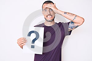 Young handsome man with tattoo holding question mark stressed and frustrated with hand on head, surprised and angry face