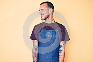 Young handsome man with tattoo wearing barber apron looking away to side with smile on face, natural expression