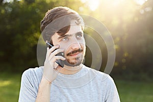 Young handsome man with thick beard and dark big eyes holding smart phone phoning his friend while standing at green background. H