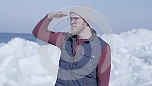 Young handsome man tourist focused on something near cold ice snowing glacier behind the winter sea. Slow motion