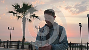 Young handsome man traveler using smartphone for book accommodation outdoor near sea coast at sunset. Caucasian guy