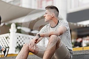 Young handsome man with trendy hairstyle in a stylish striped t-shirt in fashionable beige shorts sits on the stairs in the city