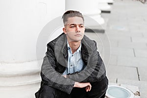 Young handsome man in a trendy plaid jacket in a stylish shirt in jeans with a fashionable hairstyle sits and enjoys a spring day