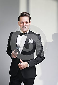 Young handsome man in a tuxedo and bow tie holding a microphone, posing over white and grey background