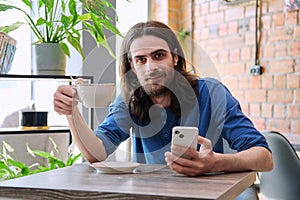 Young handsome man using smartphone, drinking cup of coffee, sitting in cafe