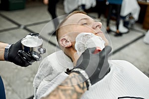 Young handsome man visiting barbershop. Cropped photo of a barber with tattooed arm applying shaving foam with brush on