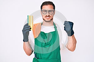 Young handsome man wearing apron holding scourer annoyed and frustrated shouting with anger, yelling crazy with anger and hand