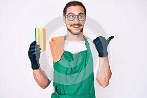 Young handsome man wearing apron holding scourer pointing thumb up to the side smiling happy with open mouth