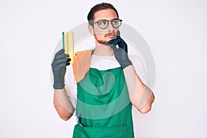 Young handsome man wearing apron holding scourer serious face thinking about question with hand on chin, thoughtful about