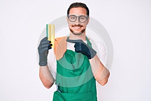 Young handsome man wearing apron holding scourer smiling happy pointing with hand and finger