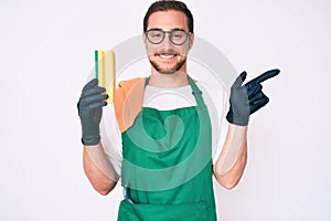 Young handsome man wearing apron holding scourer smiling happy pointing with hand and finger to the side