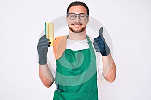 Young handsome man wearing apron holding scourer smiling happy and positive, thumb up doing excellent and approval sign