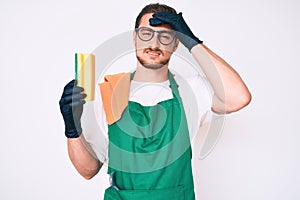 Young handsome man wearing apron holding scourer stressed and frustrated with hand on head, surprised and angry face