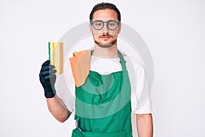Young handsome man wearing apron holding scourer thinking attitude and sober expression looking self confident