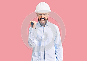 Young handsome man wearing architect hardhat angry and mad raising fist frustrated and furious while shouting with anger