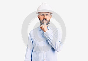 Young handsome man wearing architect hardhat asking to be quiet with finger on lips