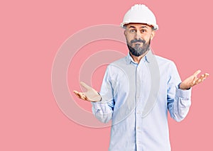 Young handsome man wearing architect hardhat clueless and confused expression with arms and hands raised