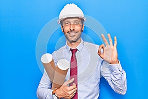Young handsome man wearing architect hardhat holding blueprint doing ok sign with fingers, smiling friendly gesturing excellent