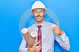 Young handsome man wearing architect hardhat holding blueprint smiling happy and positive, thumb up doing excellent and approval