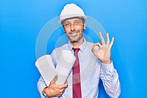 Young handsome man wearing architect hardhat holding blueprints doing ok sign with fingers, smiling friendly gesturing excellent