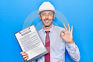 Young handsome man wearing architect hardhat holding contract document doing ok sign with fingers, smiling friendly gesturing