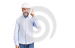 Young handsome man wearing architect hardhat pointing finger up with successful idea