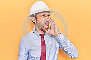 Young handsome man wearing architect hardhat shouting and screaming loud to side with hand on mouth