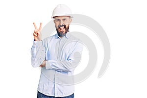 Young handsome man wearing architect hardhat smiling with happy face winking at the camera doing victory sign