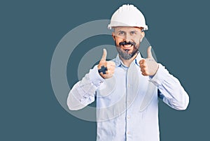 Young handsome man wearing architect hardhat success sign doing positive gesture with hand, thumbs up smiling and happy