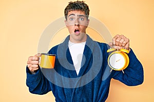 Young handsome man wearing bathrobe holding cup of coffee and alarm clock afraid and shocked with surprise and amazed expression,