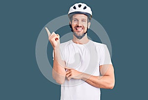 Young handsome man wearing bike helmet with a big smile on face, pointing with hand and finger to the side looking at the camera