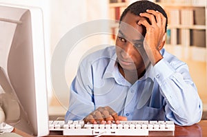 Young handsome man wearing blue office shirt sitting by computer leaning onto desk while typing and looking uninspired photo