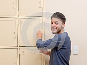 Young handsome man wearing blue sweater smiling and opening metal locker door, rack of lockers stacked