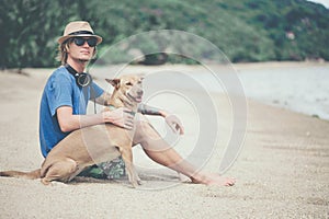 Young handsome man wearing blue t-shirt, hat and sunglasses, sitting on the beach with the dog