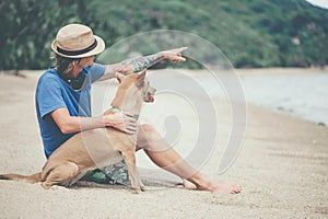 Young handsome man wearing blue t-shirt, hat and sunglasses, sitting on the beach with the dog