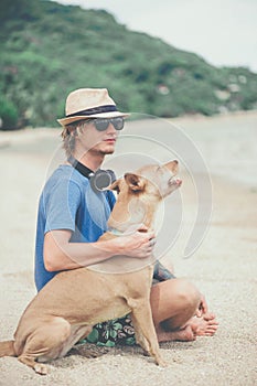 Young handsome man wearing blue t-shirt, hat and sunglasses, sitting on the beach with the dog in Thailand