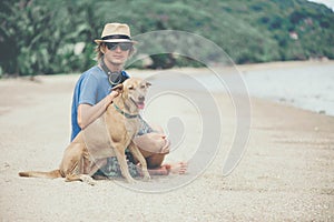 Young handsome man wearing blue t-shirt, hat and sunglasses, sitting on the beach with the dog in Thailand