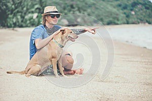 Young handsome man wearing blue t-shirt, hat and sunglasses, sitting on the beach with the dog