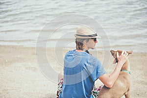 Young handsome man wearing blue t-shirt, hat and sunglasses, sitting on the beach with the dog in Thailand