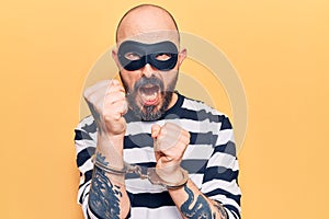 Young handsome man wearing burglar mask and handcuffs annoyed and frustrated shouting with anger, yelling crazy with anger and