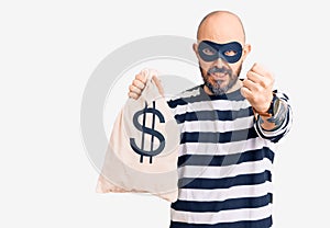 Young handsome man wearing burglar mask holding money bag with dollar symbol annoyed and frustrated shouting with anger, yelling