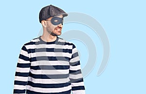 Young handsome man wearing burglar mask looking away to side with smile on face, natural expression